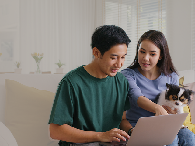 Homem de camiseta verde mexendo no notebook ao lado de mulher de camiseta roxa apontando para o notebook com cachorro em seu colo